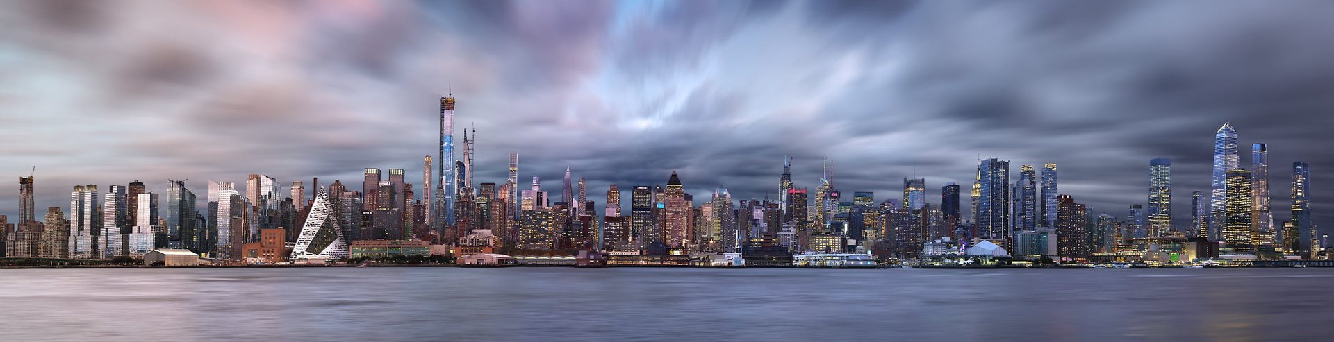 A panorama of a big city. It transitions from sunset to nighttime from left to right. There are lots of skyscrapers across a waterfront.
